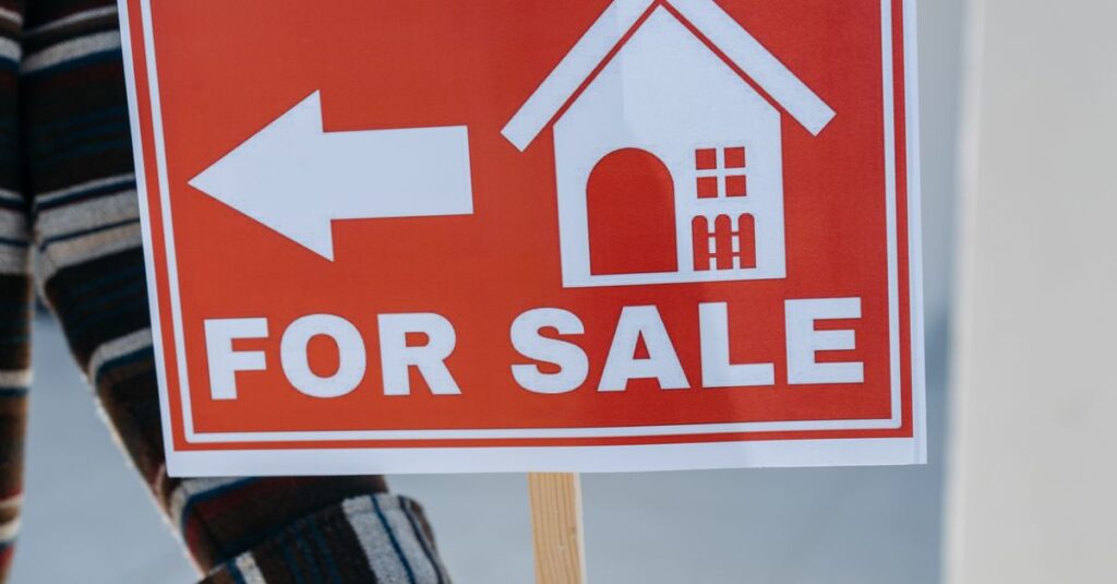 Close-up of a hand holding a 'For Sale' real estate sign with a house icon outdoors.