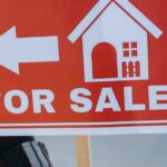 Close-up of a hand holding a 'For Sale' real estate sign with a house icon outdoors.